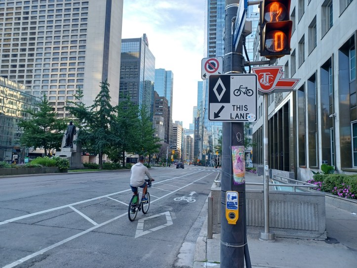 Another bike lane in the downtown core of the city. This one is not elevated. The street sign clearly states where the bikes should go. The TTC sign on the red background in the back marks a subway entry point.
